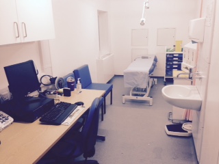 completed room. Showing clinicians desk at the front, examination bed in, chairs to one side and cabinets on the other.